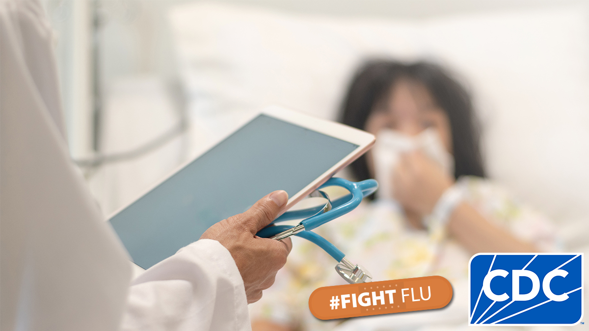 A doctor holding a tablet and a patient in a hospital bed.