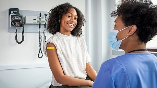 A healthcare provider wearing a mask and chats with a smiling patient with a bandage on their upper arm.