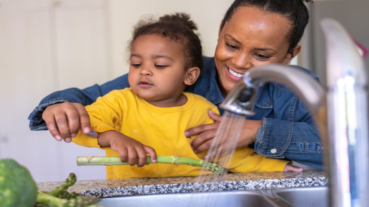 Parent and child preparing to wash produce under running water coming out of a sink faucet