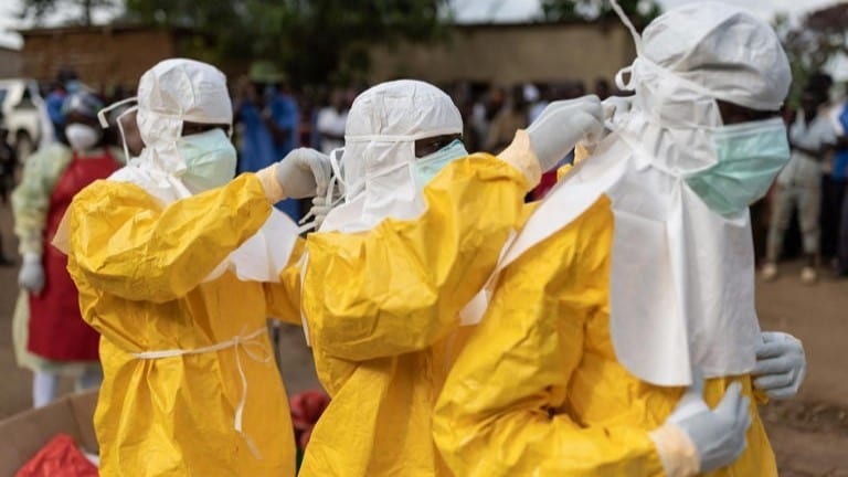 Three men wearing yellow hazmat suits assisting each other with putting on PPE