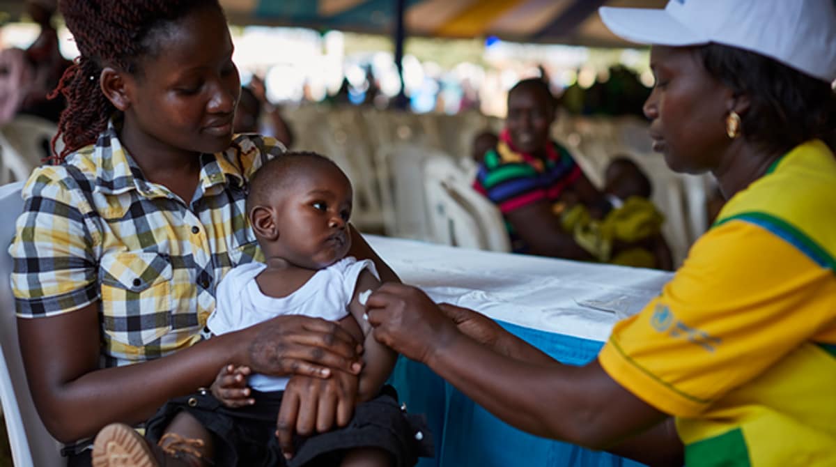 A woman in a yellow and green uniform administers a malaria vaccine to a baby held by his mother.