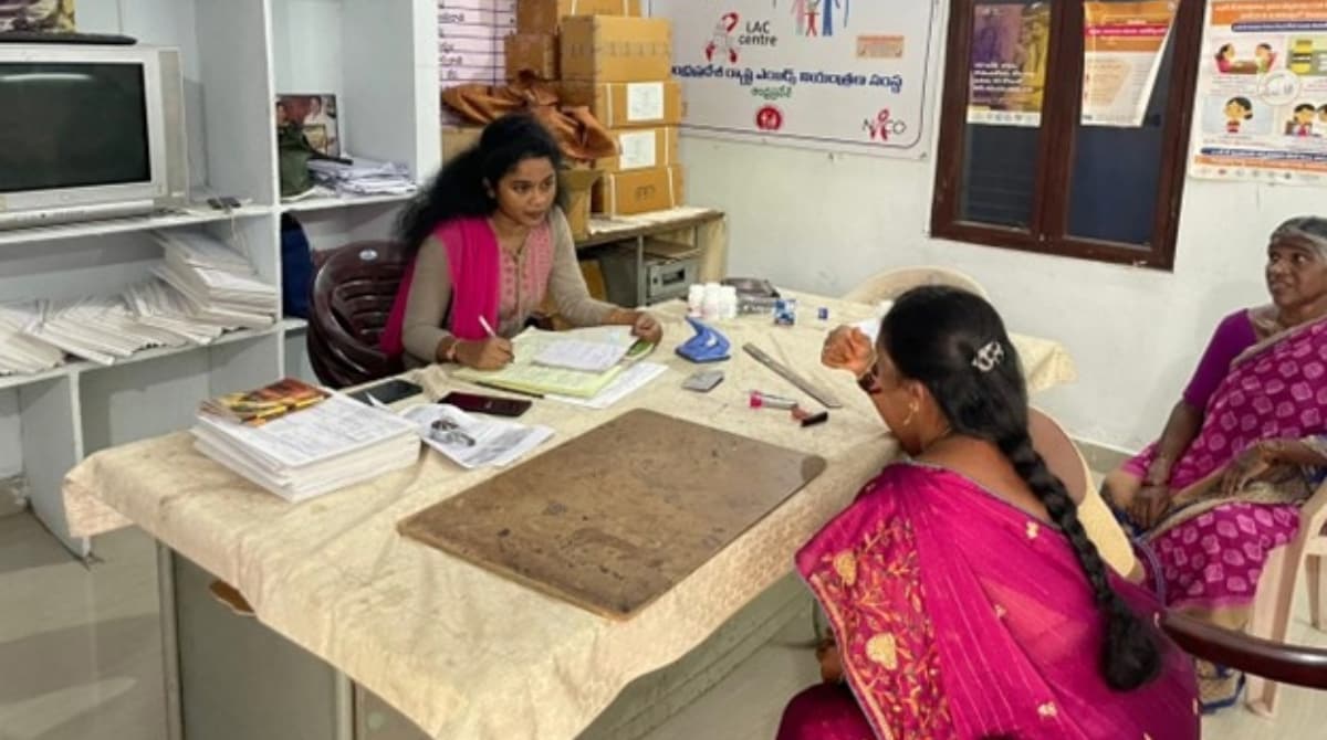 A woman in a health clinic sits at a table while she fills paperwork. She is engaging with two other women seating across the table. The room contanains various files and suplies around.