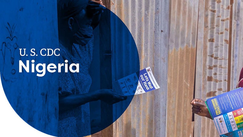 People handing out health informational flyers near a corrugated metal wall, labeled "U.S. CDC Nigeria."