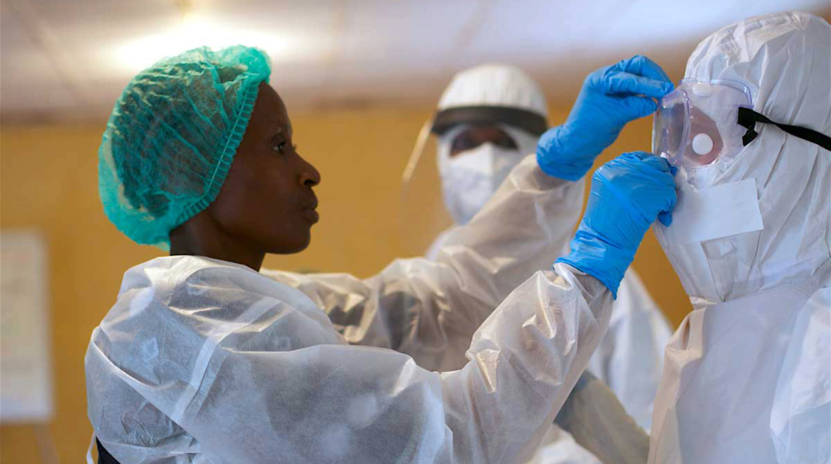 A healthcare worker in a green hairnet and blue gloves helps another person in a white protective suit adjust their safety goggles.