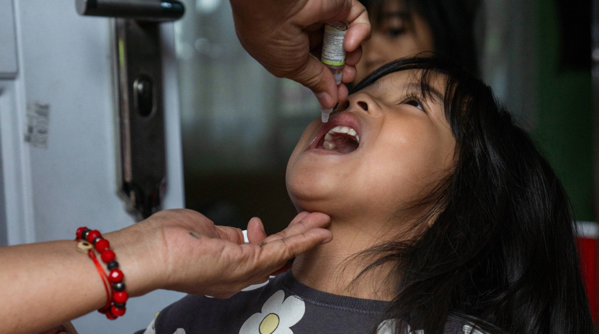 A toddler looks up as a healthcare worker administers oral vaccine drops into their mouth from a small vial.