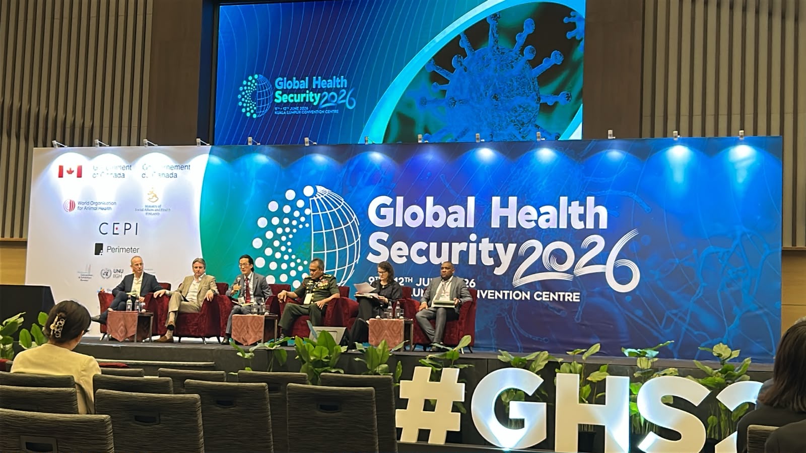 Panel of six speakers on a stage with a "Global Health Security 2026" blue banner background behind them.