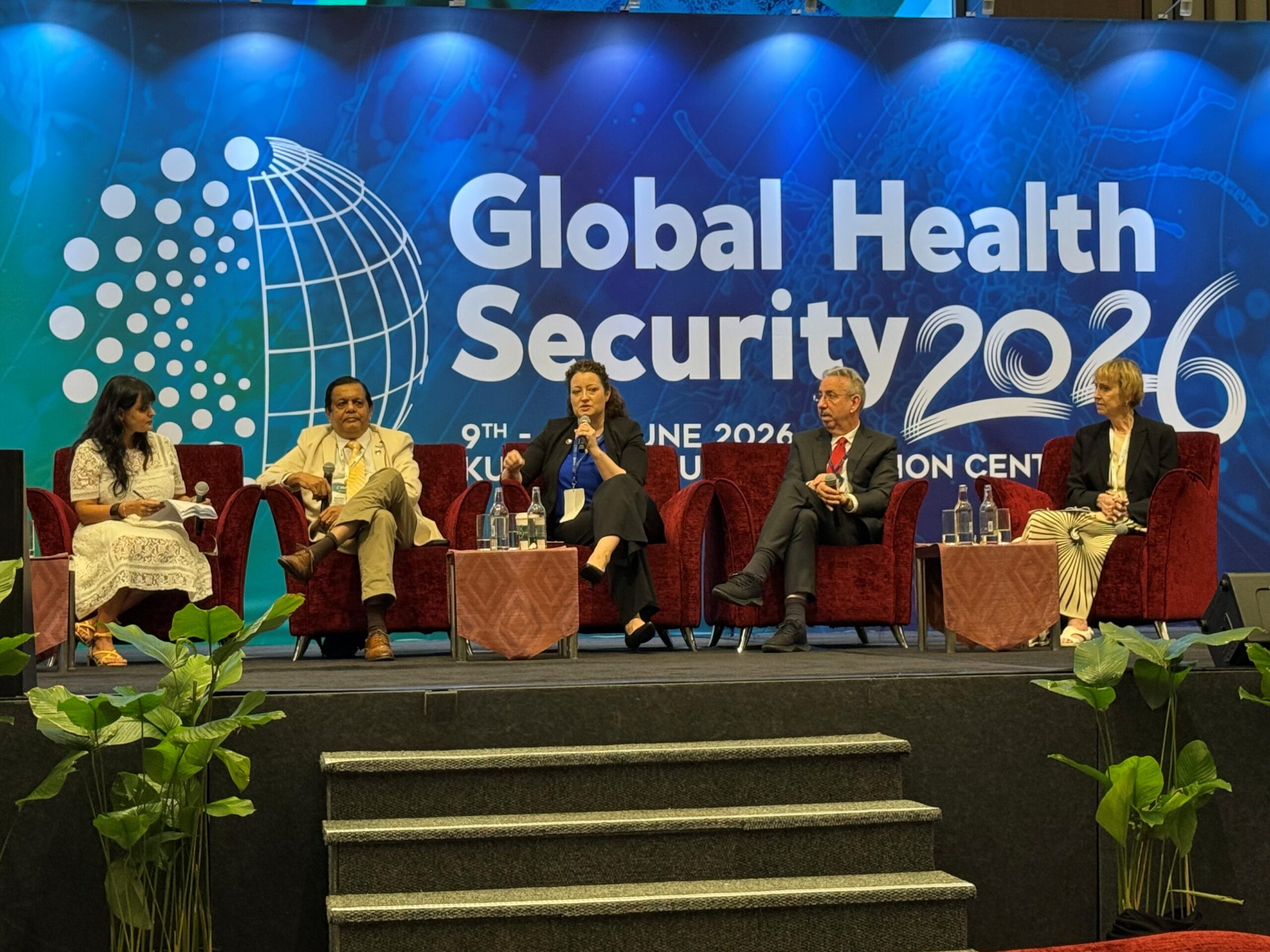 Panel of 5 people speaking with a "Global Health Security 2026" blue banner behind them. They are all in a stage and a woman hold a microphone.