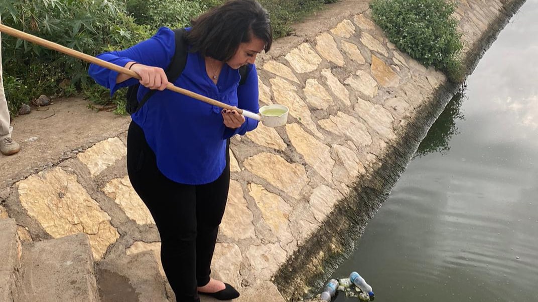 Woman in blue shirt and black pants standing near body of water lifts net out and inspects contents.