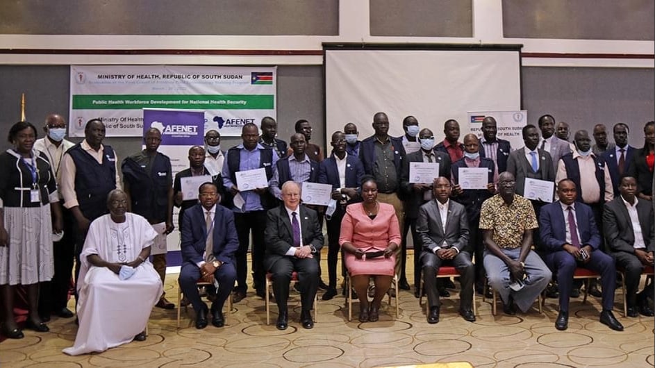 A photo of over 30 people standing or sitting closely together for a group photo. Some people are holding up certificates. A banner behind them reads, "Ministry of Health, Republic of South Sudan, Public Health Workforce Development for National Health Security."
