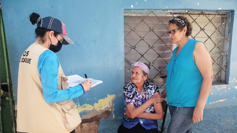 A masked health worker holds a notebook and speaks with two local women about the importance and availability of getting vaccines.