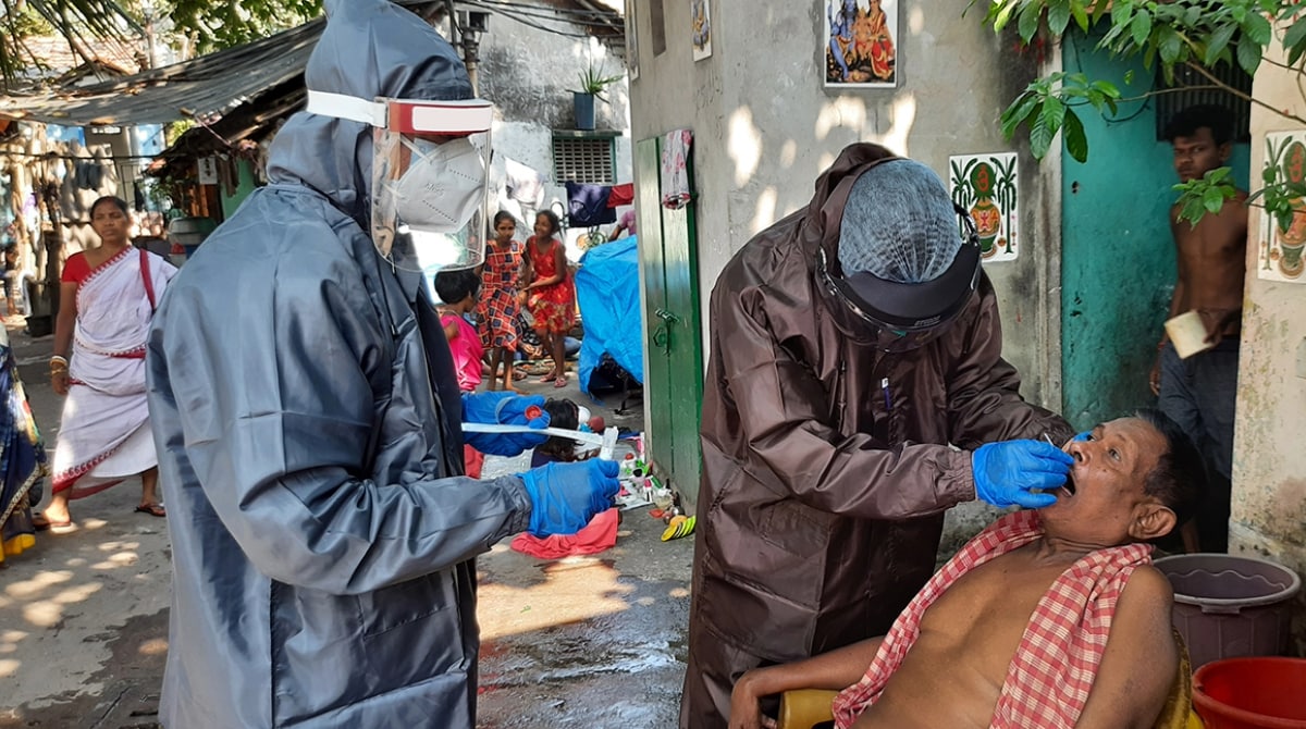 Two nurses in protective gear swab a man's mouth in an outdoor setting.