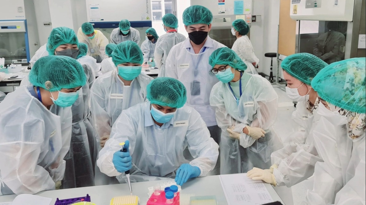 A group of scientists, in personal protective equipment, huddle around a table, in a lab, to watch one person who is holding a pipette over a desk.