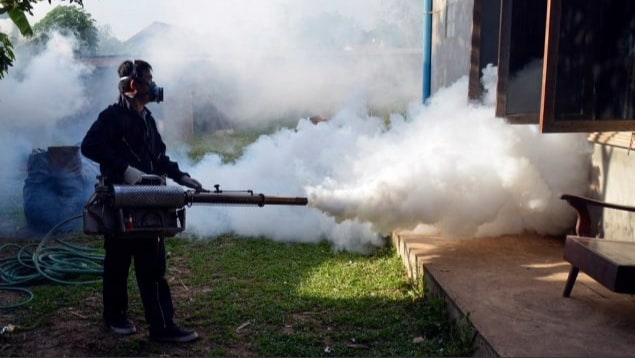 Person wearing a respirator is spraying pesticides along the outside of a dwelling.