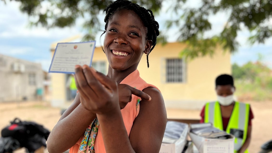 A teen girl in a coral top stands outside and holds up her vaccine card as she points to her arm where she just received a COVID-19 vaccine.