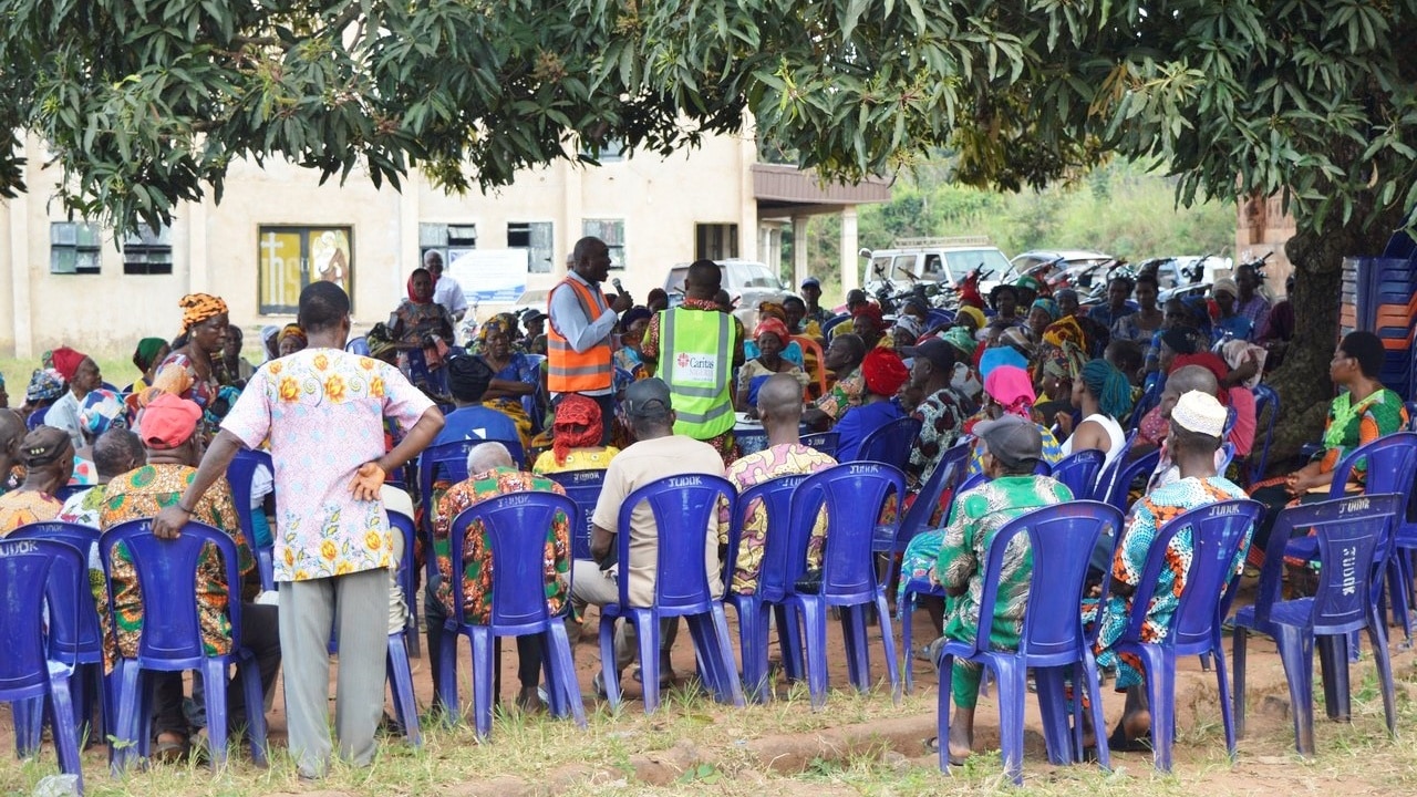 A large group of individuals sit outside in chairs. A man with a microphone stands in the middle to share a message about COVID-19.