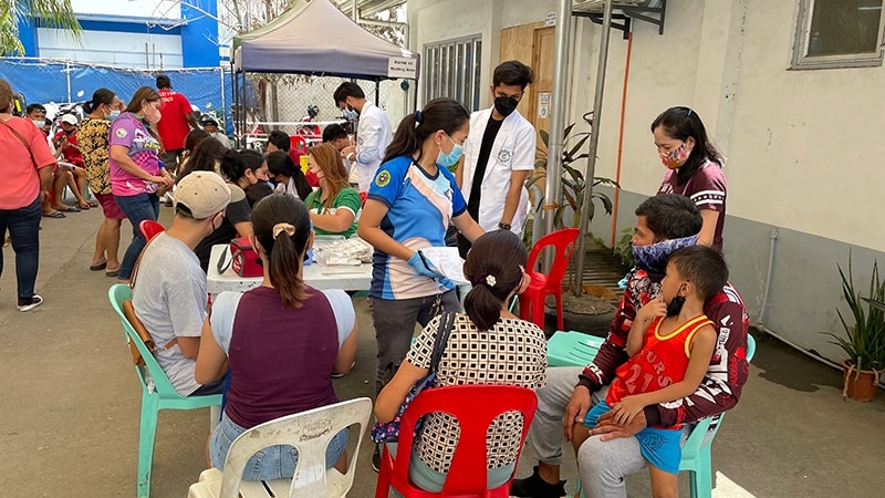 Many people with face masks covering their nose and mouth are standing or sitting outdoors near a table and tent.