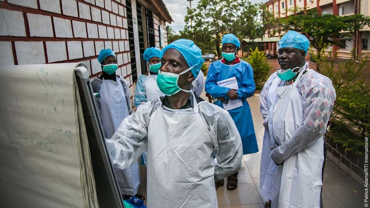 A health worker writes on a flip board. Four other health workers stand behind him. All are wearing full personal protective equipment.