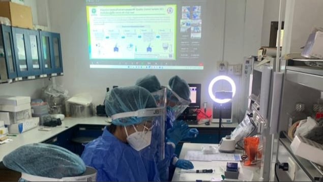 Laboratorians wearing personal protective equipment sit side-by-side in a laboratory. Pipettes and other lab supplies are on the desk in front of them, and a presentation slide is projected onto the wall behind them.