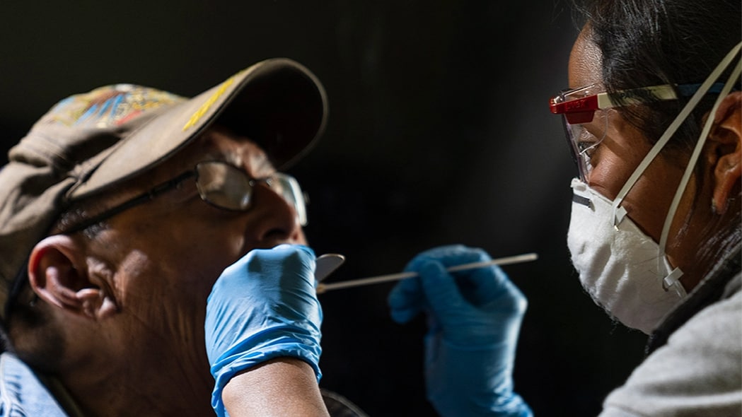 A person wearing a mask and gloves holds a swab in another person's mouth to collect an oral sample.