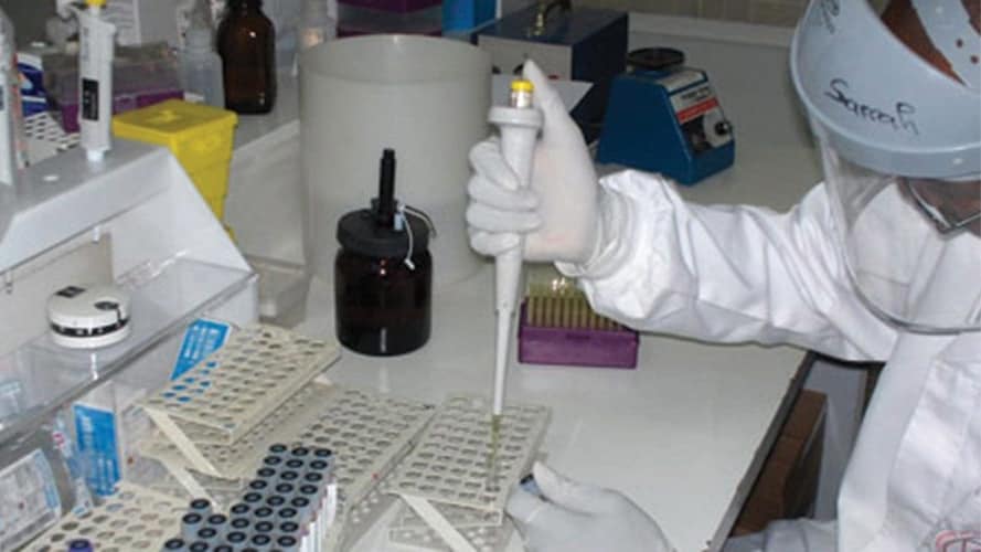 Photo of laboratorian wearing personal protective equipment and holding a pipette over a test tube sitting in a test tube rack.