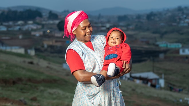 Woman stands outside smiling and holding her baby.