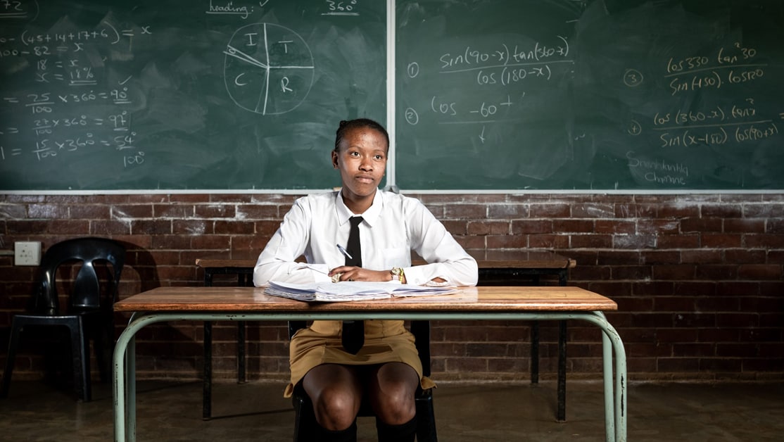 A girl in a white shirt sits at a desk with a chalkboard behind her.