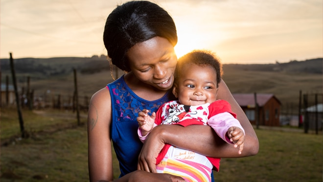 A mother stands in a field smiling and holding her baby girl.