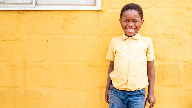 A smiling 11-year-old boy wearing a yellow shirt standing against a yellow wall