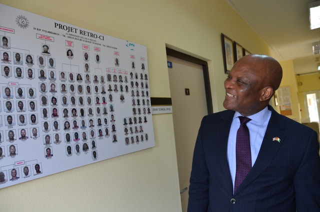 Dr. John Nkengasong stands in a hallway smiling while looking at a poster of individuals working for Project Retro-CI.
