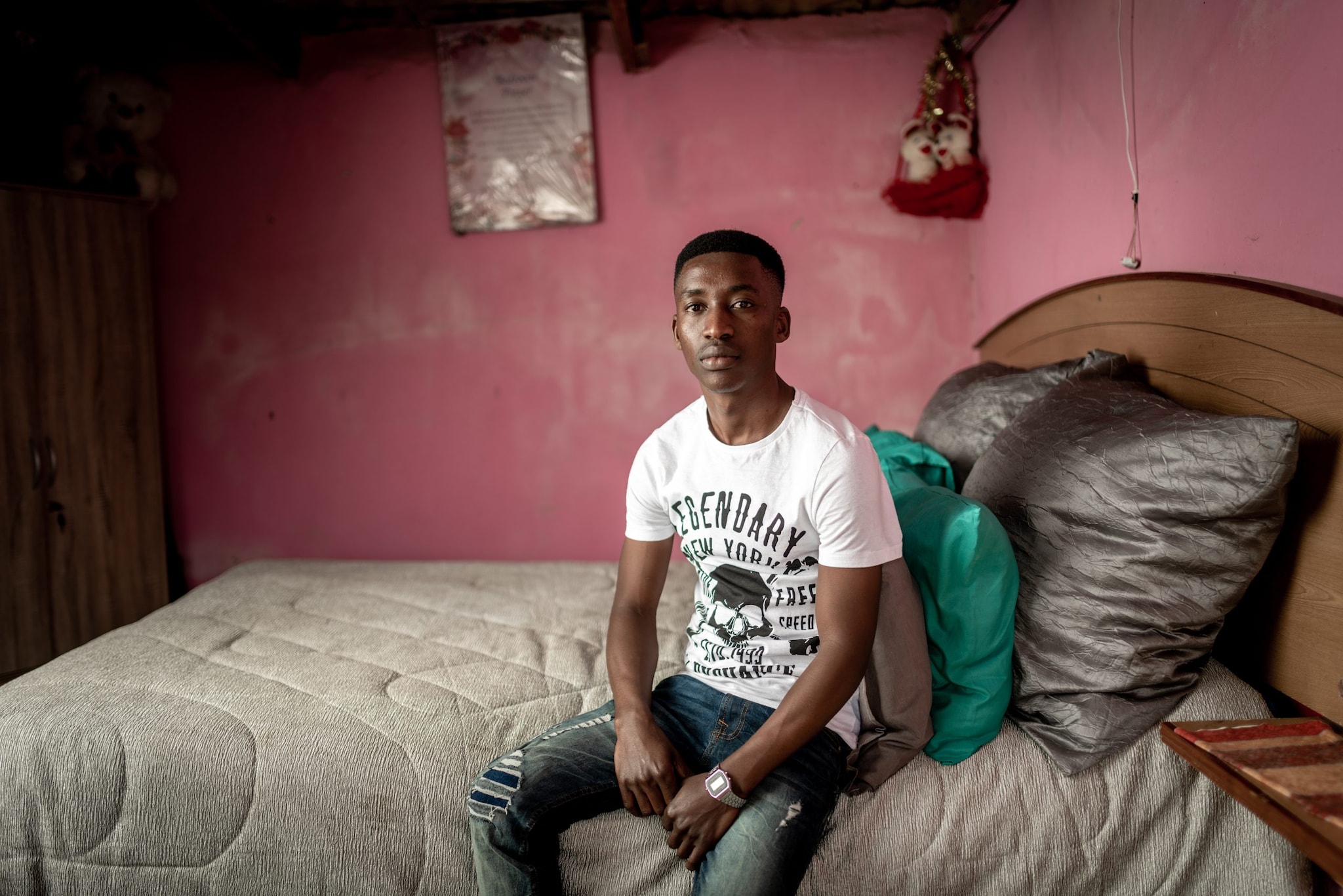 Young man in white shirt and jeans sits on bed.