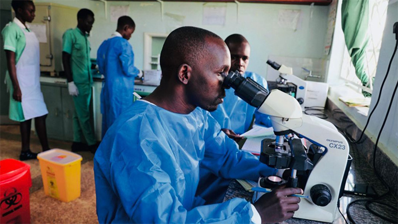 Man in blue coat examines through microscope.