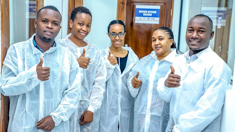 Group of 5 people in lab coats give thumbs up. They stand in front of an "HIVDR Store Room" signage.