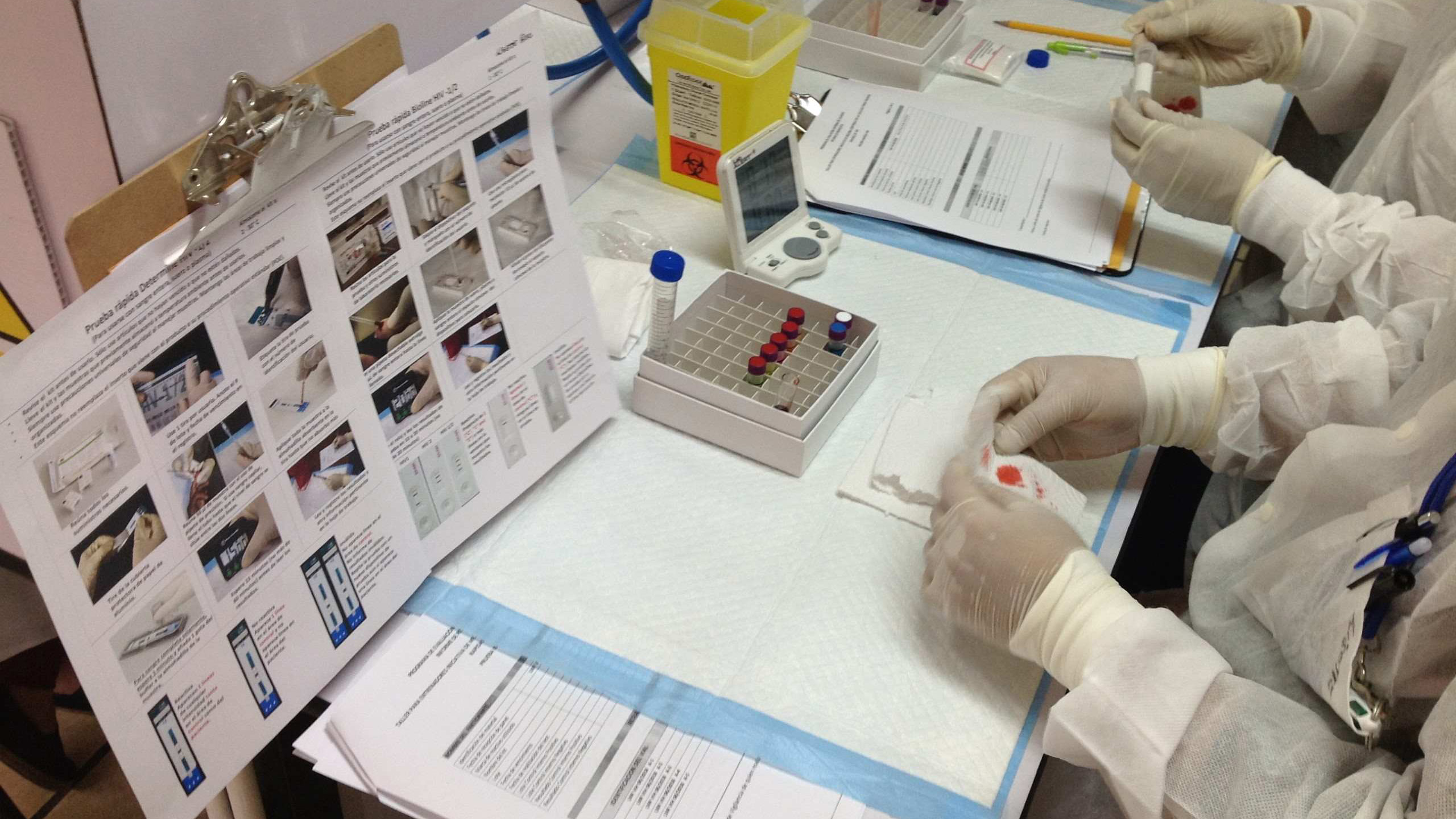 Lab staff in protective gear prepare blood samples using testing supplies and documentation.