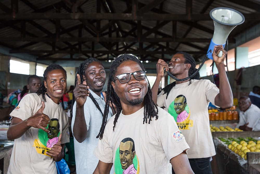 A group of artists in matching shirts gather and smile at a market in Mozambique.