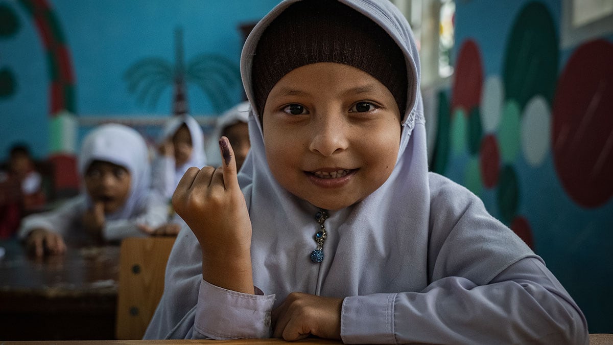 A young girl sitting at a school desk smiles while she holds up her pinky finger, which has been marked to show she received a polio vaccine.