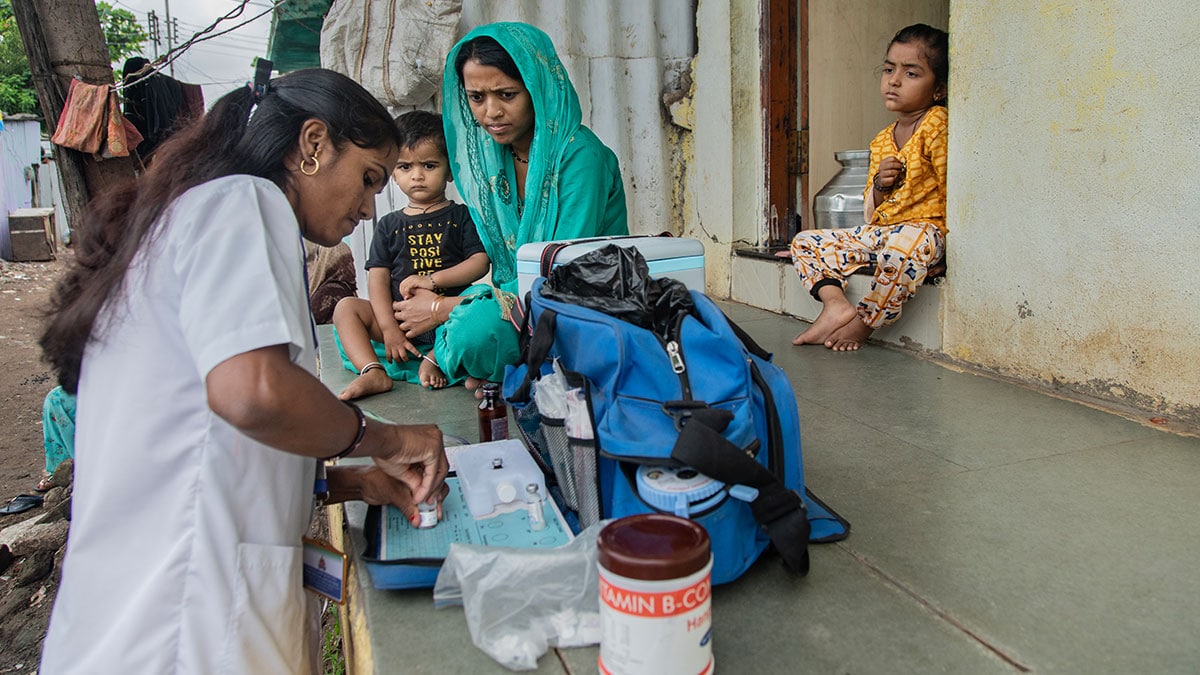 A woman in a white coat twists the lid of a vaccine vial on a porch, where a woman and two children are sitting and where there is a blue bag and a container labeled "vitamin."