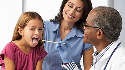 A doctor examines a young girl’s throat using a tongue suppressor. Smiling mother in background.