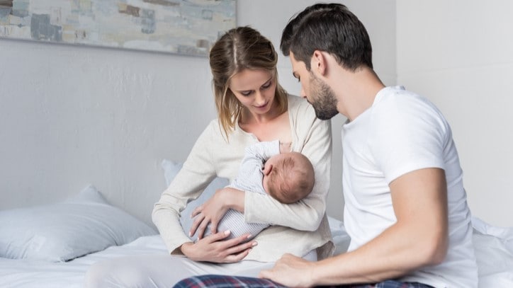 Mother sitting on a bed breastfeeding an infant while the father offers support.