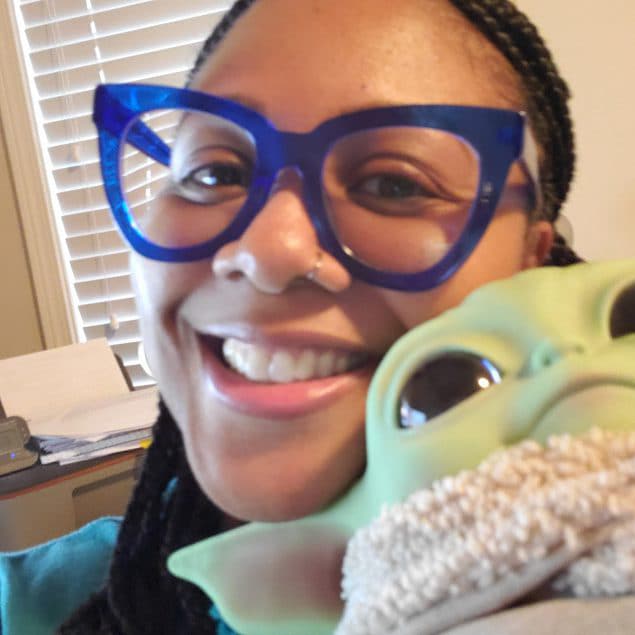 Melinda Jordan wearing blue glasses and holding a doll from the Star Wars franchise, Grogu.