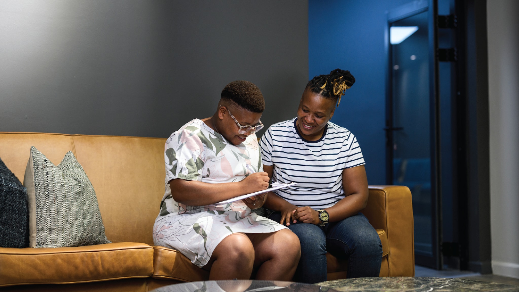 Two people sitting side-by-side on a couch as one of them fills out forms on a clipboard.