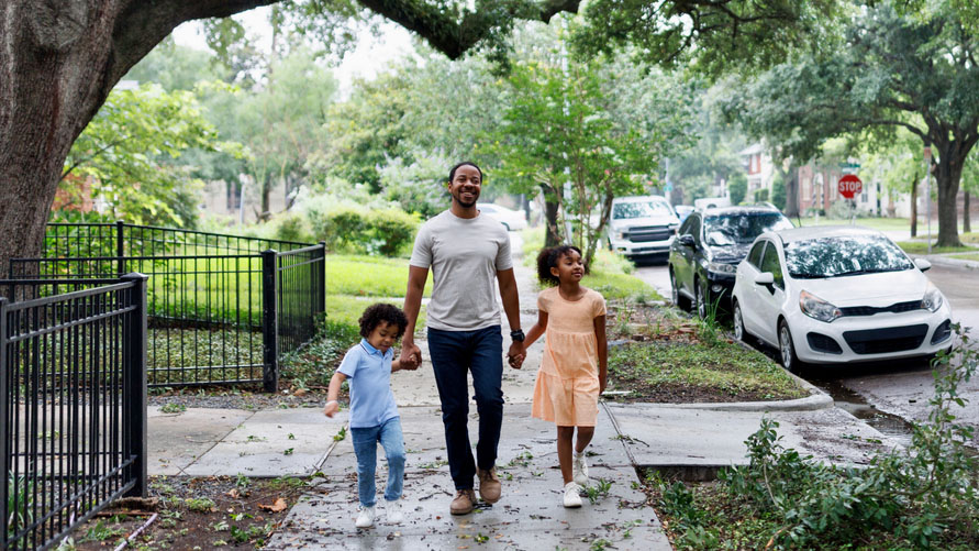 A dad is holding the hands of two kids while walking on a sidewalk