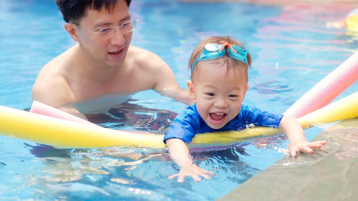 Un padre con su hijo pequeño en la piscina. El padre sostiene al niño mientras este nada con un flotador para piscinas.
