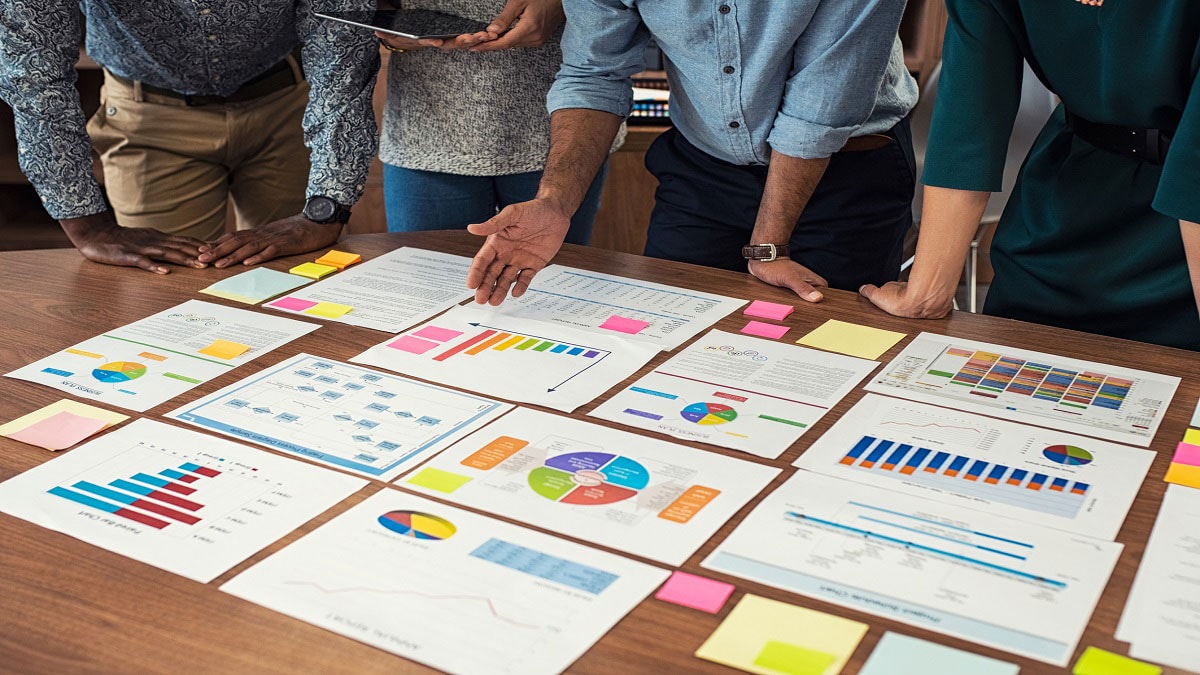 Printed paper with different graphs and charts laid out on a table, with coworkers standing around the table.