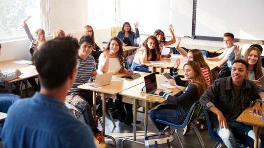 A male teacher in front of a class of students who are raising their hands.