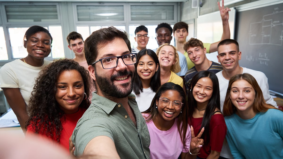 A male teacher taking selfie with a group of multi racial high school students in front of classroom.