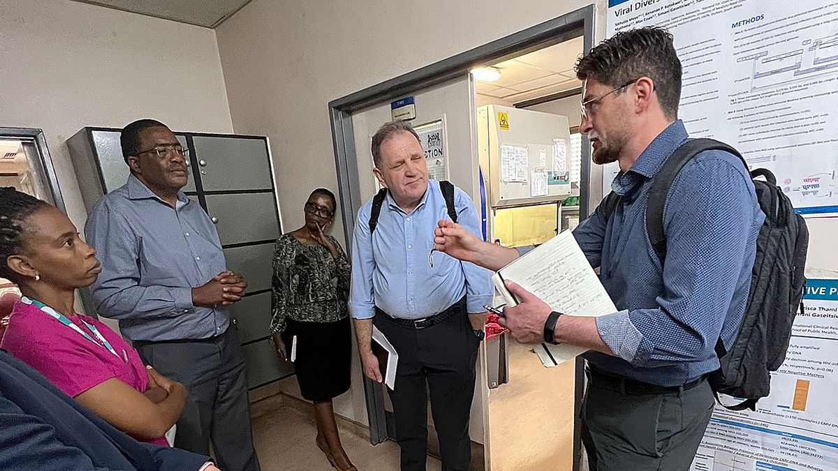 Five people discuss in a lab setting, with a speaker gesturing toward a scientific poster on the wall