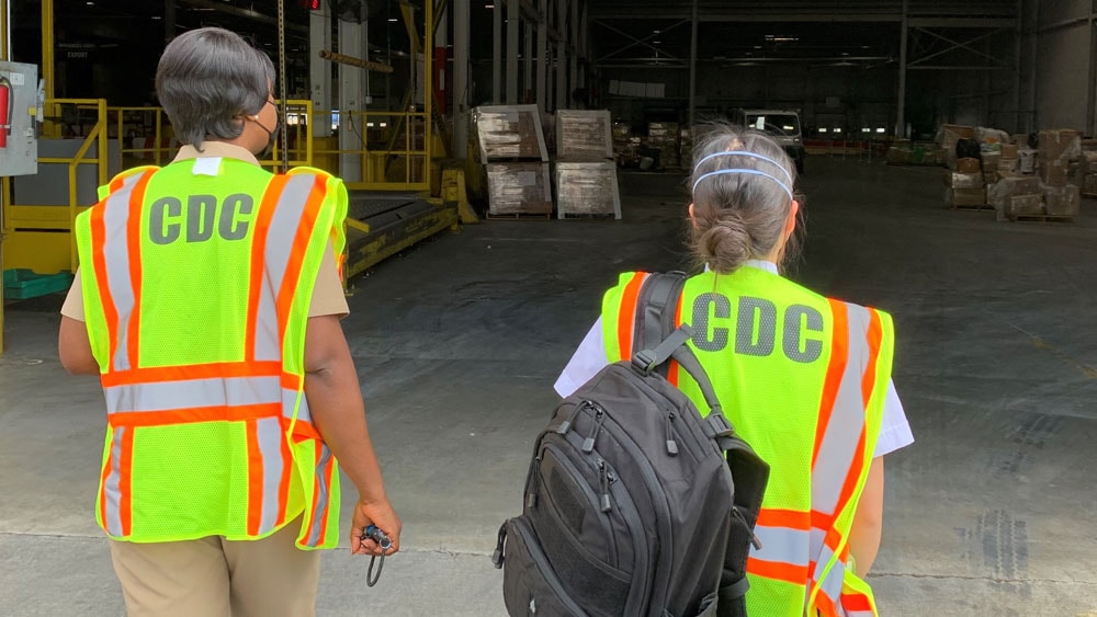 Workers wearing CDC vests walk toward a warehouse.