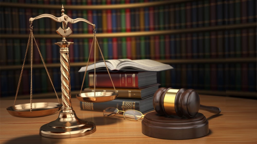 Stack of books, gavel, and scales of justice on a table in front of a bookshelf.