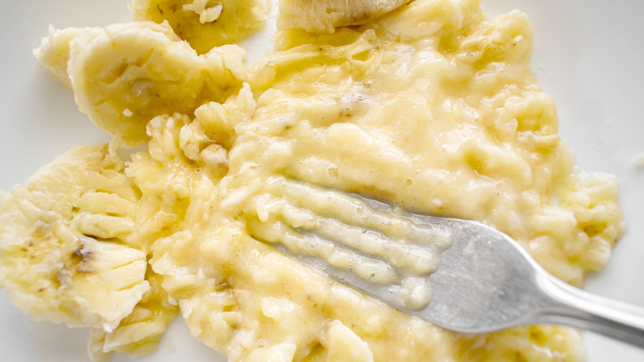Ripe banana being mashed with a fork on a white plate.