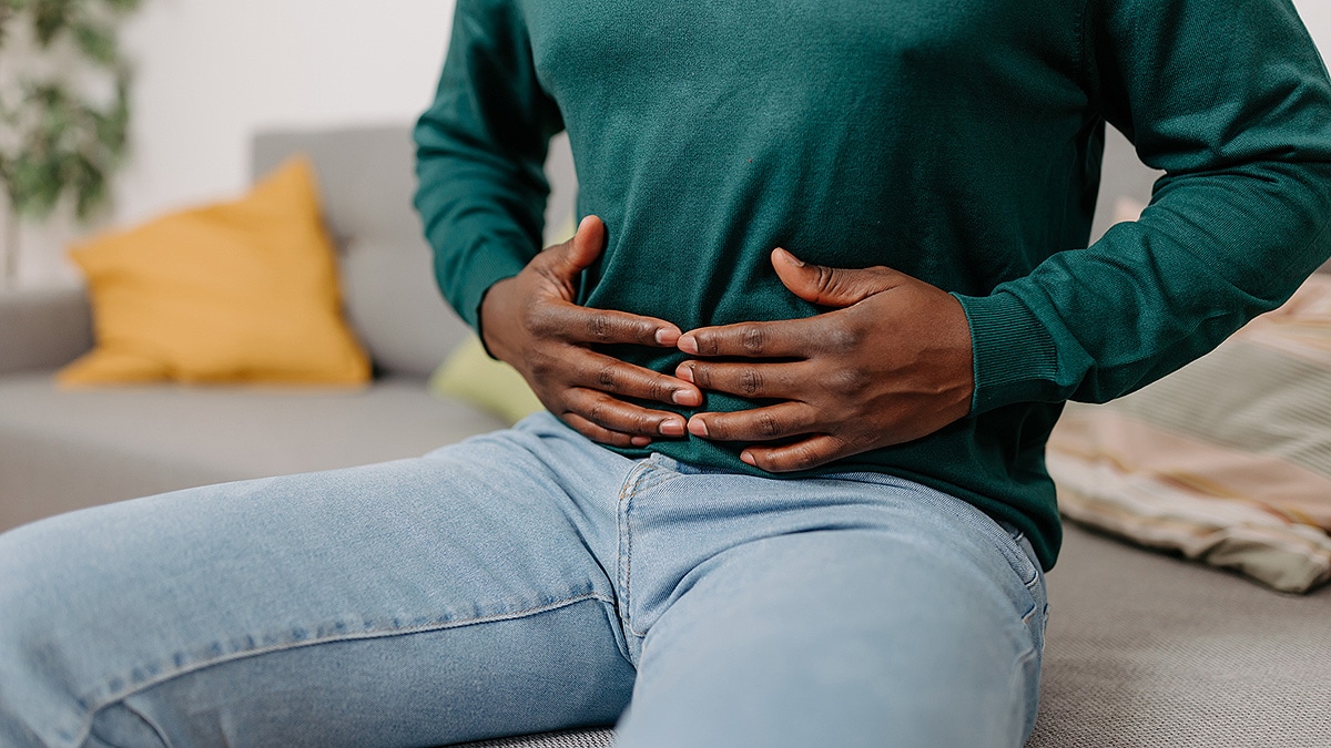 A young African American man sits on a sofa, holding his stomach from digestive pain.